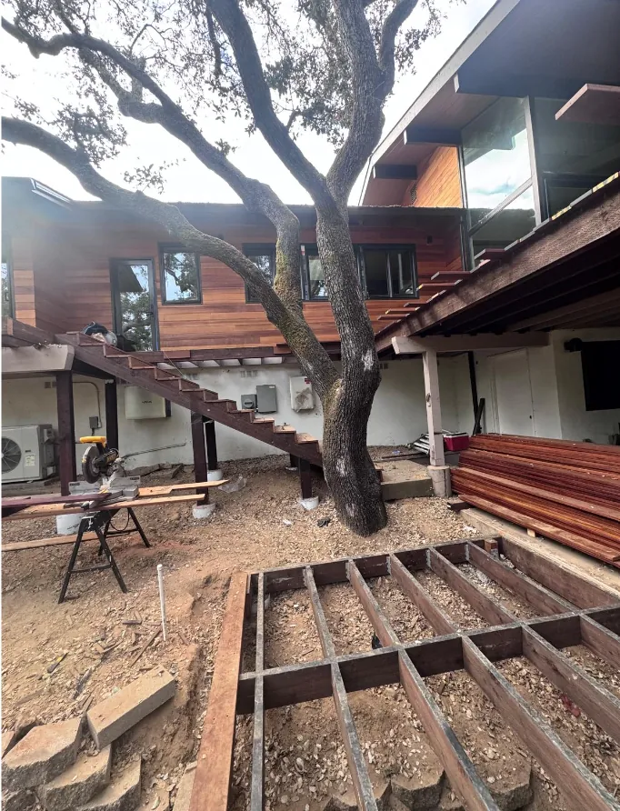 Deck joist framing laid out on concrete piers over a graded pad beside a mature oak, in front of a wood-clad hillside home with an exterior wood staircase, a miter saw station and stacked hardwood decking.