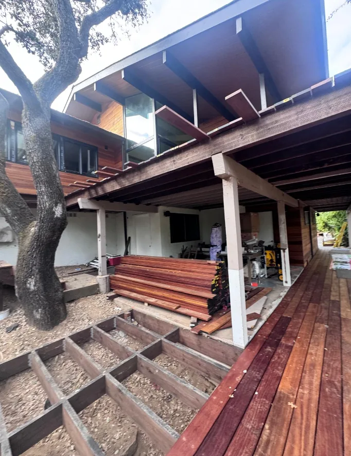 Deck joist framing laid out over a gravel pad beside a stack of hardwood decking boards, beneath the posts and beams of an elevated deck with a finished hardwood walkway alongside.