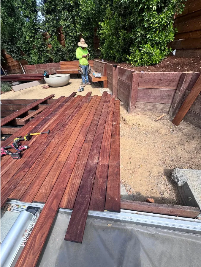 Hardwood deck boards being laid across deck framing in a back yard, with stacked timber retaining walls, a built-in wood bench and a round concrete bowl behind.