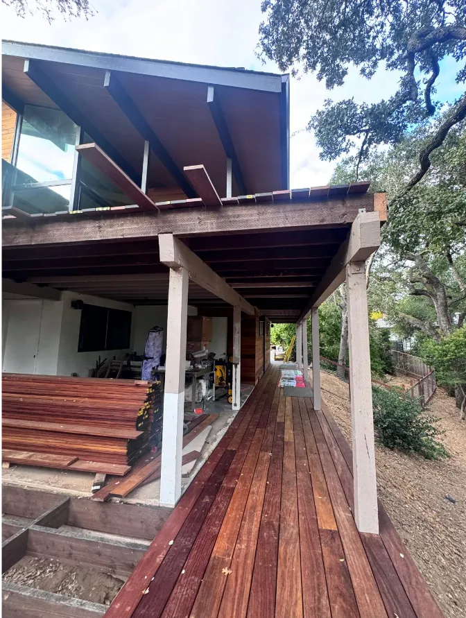 Finished hardwood walkway running beneath the beam and rafters of an elevated deck on a hillside home, with bundles of hardwood decking stacked in the covered work area alongside.