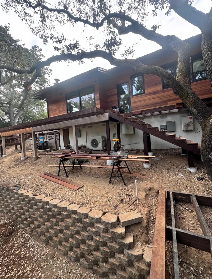 Deck and exterior staircase under construction on a wood-clad two-story hillside home, with a segmental concrete block retaining wall below and lumber laid across sawhorses at the cut station.