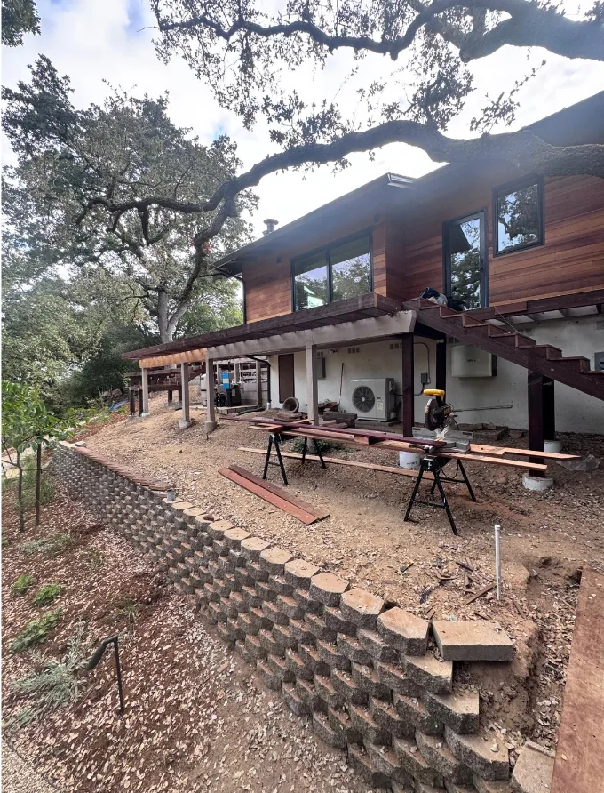 Wood-clad hillside home with an elevated deck across the upper floor, an open exterior staircase down to grade and a segmental block retaining wall stepping down the slope, beneath a spreading oak.