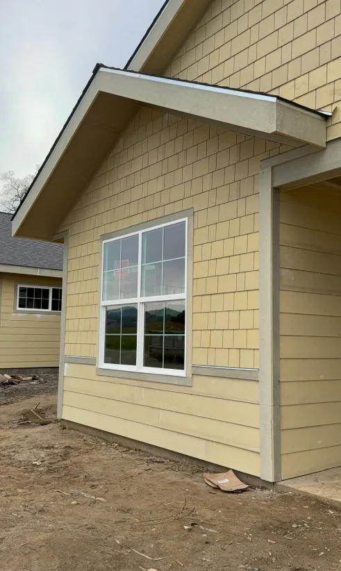 New home exterior clad in yellow shingle siding above lap siding, with boxed eaves, white corner trim and a newly installed window.