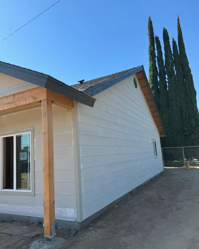 Completed new home with white lap siding and a covered porch supported by a natural wood post, with Italian cypress alongside.