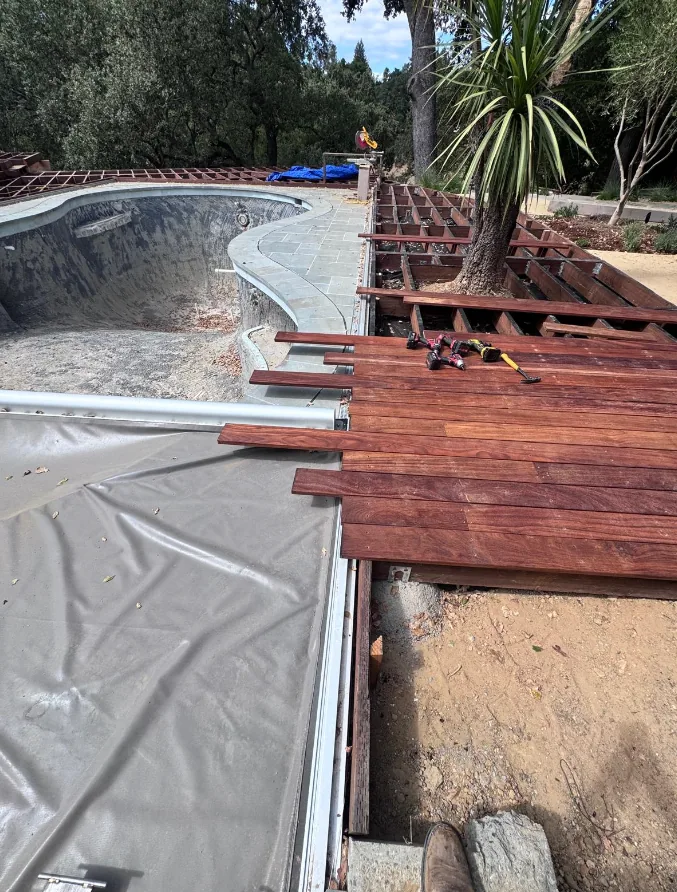 Hardwood deck boards being installed over joist framing beside an empty pool shell under refinishing, with curved stone coping and cordless tools resting on the new decking.