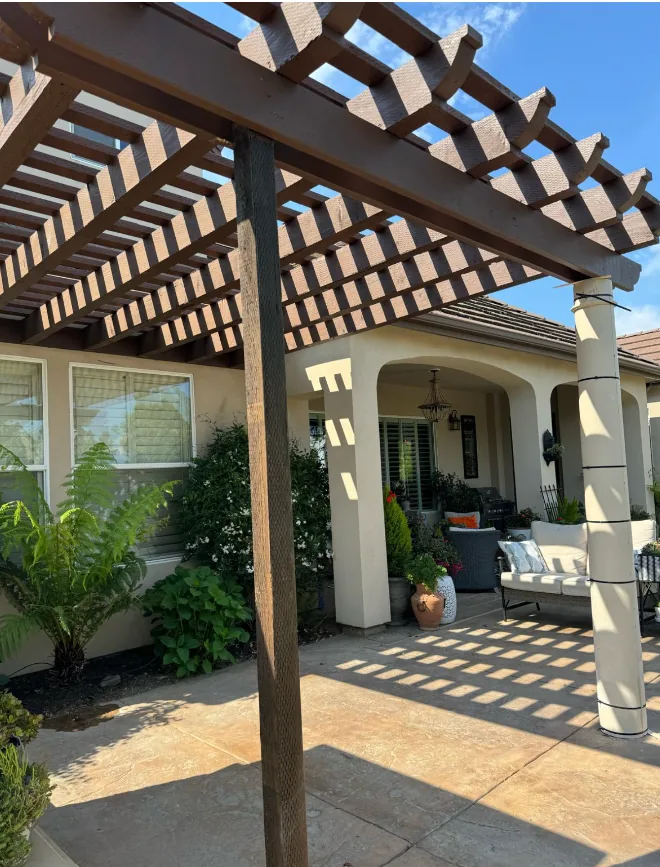Stained wood pergola patio cover casting striped shade over a stamped concrete patio beside a stucco home's covered entry.